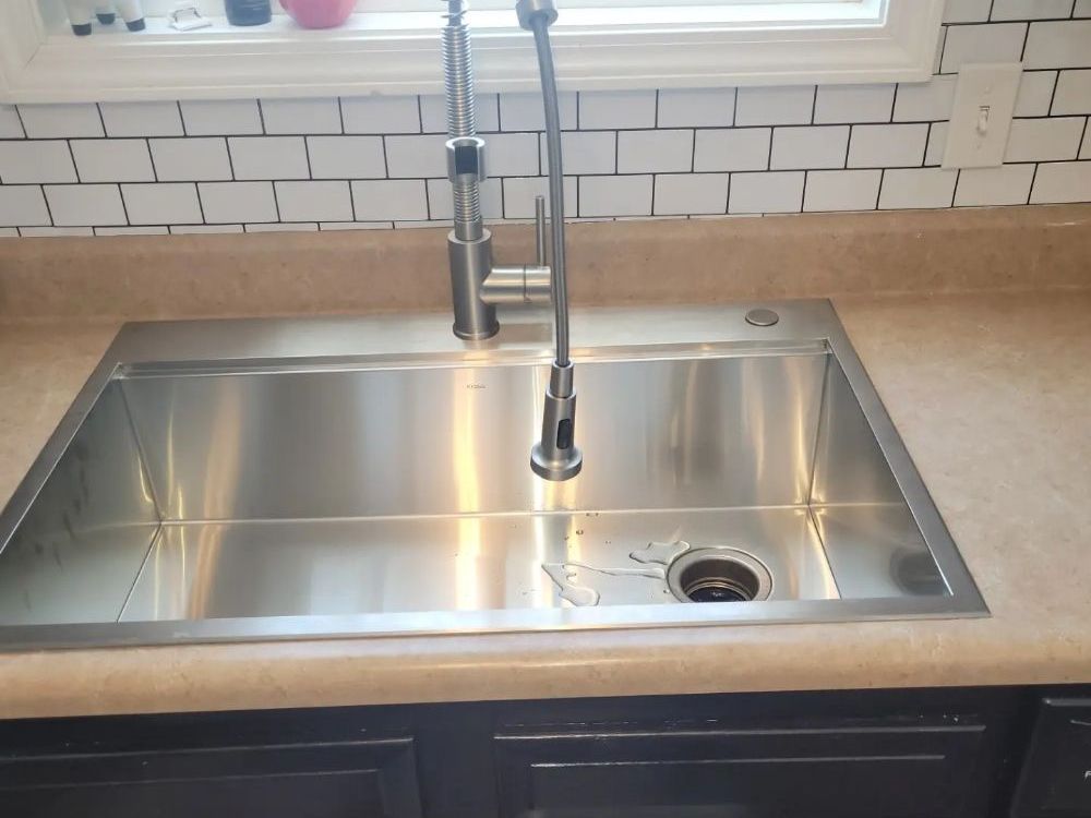 A new stainless steel kitchen sink with a tall spring faucet, set in a counter below a window, with white subway tile behind and dark cabinets underneath.