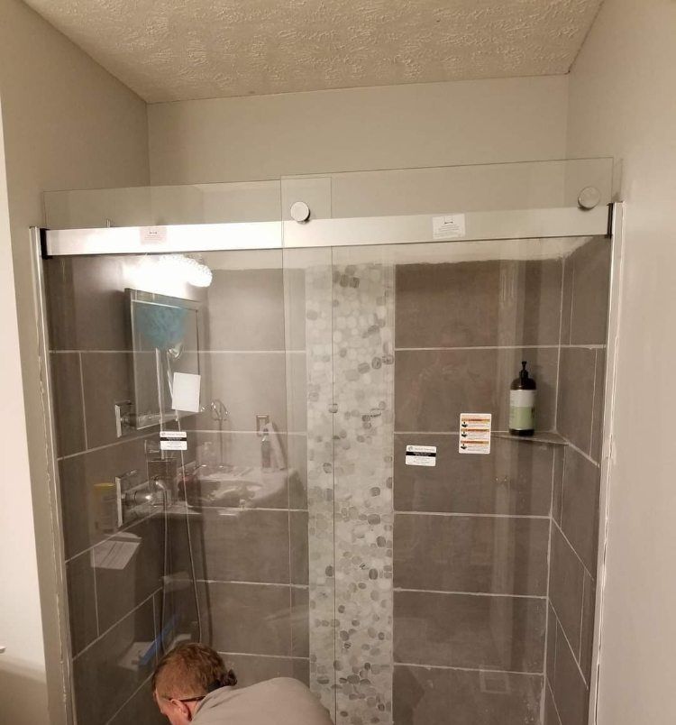 A worker kneeling to fit a frameless glass shower enclosure in a bathroom tiled in grey stone-effect tile.